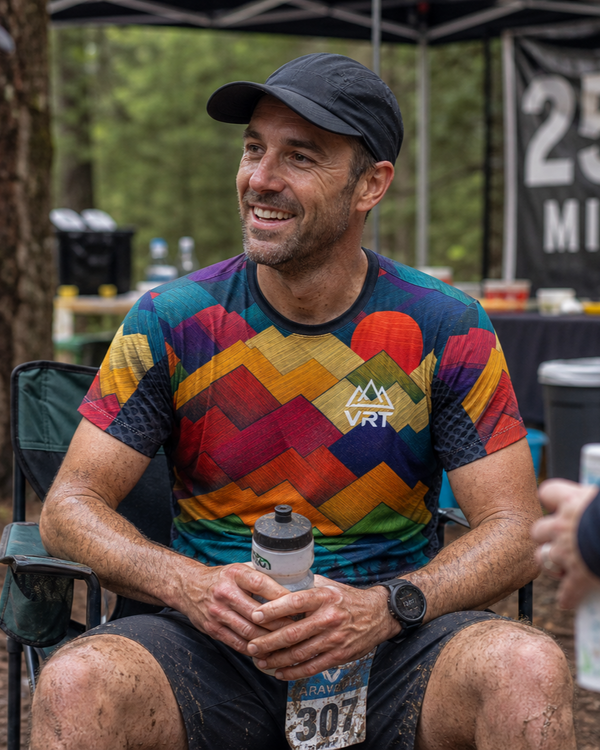 Man wearing a vrt rising peaks running shirt with 'VRT' logo, sitting outdoors at an event.